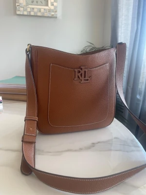Brun axelväska från Ralph Lauren - Snygg brun axelväska i läder från Ralph Lauren med justerbar rem och RL-logga framtill. Väskan har en praktisk ytterficka och stilren design med synliga sömmar. Perfekt för dig som gillar klassiska accessoarer med en modern touch.