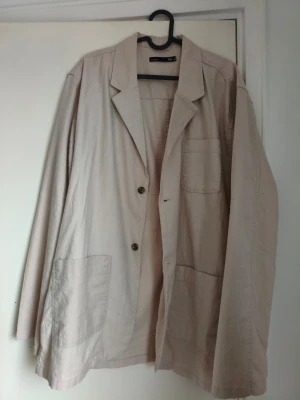 Beige linne kavaj  - Stilren beige kavaj från Lager 157 med klassisk krage, tre framfickor och två knappar framtill. Långärmad modell i lätt bomullsmaterial som passar perfekt till en avslappnad men snygg look.