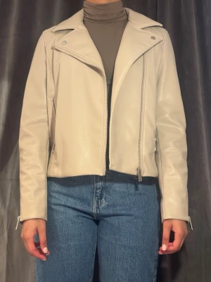 Beige skinnjacka med dragkedjor - Snygg beige skinnjacka med klassisk bikerkrage och silvriga dragkedjor framtill och vid ärmsluten. Jackan är oanvänd.