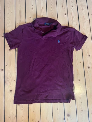 Ralph Lauren piké - Snygg vinröd pikétröja från Polo Ralph Lauren i slim fit-modell. Tröjan har klassisk krage, korta ärmar och den ikoniska broderade loggan på bröstet. Perfekt för en stilren och avslappnad look.