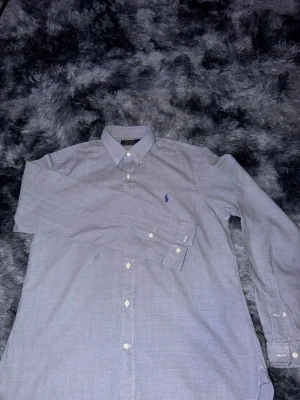 Rutig skjorta från Polo Ralph Lauren - Klassisk rutig skjorta från Polo Ralph Lauren i ljusblått med diskret rutmönster och broderad logga på bröstet. Skjortan har lång ärm, knappar framtill och button-down krage. Perfekt för en stilren look.