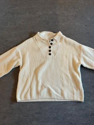 Beige/vit stickad tröja med krage - Snygg beige/vit stickad tröja med krage och fyra svarta knappar framtill. Tröjan har lång ärm och ribbad struktur, perfekt för lager-på-lager. Enkel och stilren design som passar till det mesta.