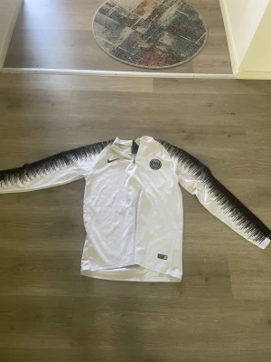 Psg tröja - Snygg vit långärmad träningströja från Nike med PSG-logga på bröstet. Ärmarna har svart grafiskt mönster som ger en cool kontrast. Perfekt för fotbollsträning eller sportiga aktiviteter. Materialet är lätt och andas bra.