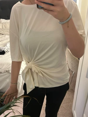Vit topp med knyt i midjan - Säljer en stilren vit topp med snygg knytning i midjan. Toppen har en lös passform och är tillverkad i ett mjukt, tunt material som känns skönt mot huden. Perfekt att styla med både jeans och kjol.