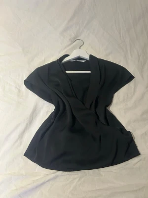 Svart omlottopp från MNG - Snygg svart omlottopp från MNG med korta ärmar och djup v-ringning. Toppen har en elegant och flowy passform som ger en stilren look. Perfekt att styla med jeans eller kjol för en trendig outfit.