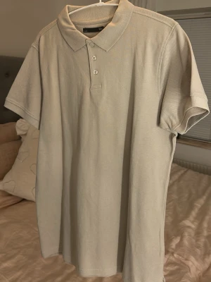 Beige pikétröja från 157 - En stilren beige pikétröja från 157 med klassisk krage och tre knappar framtill. Tillverkad i mjuk bomull och har korta ärmar. Enkel och clean design som funkar till det mesta.