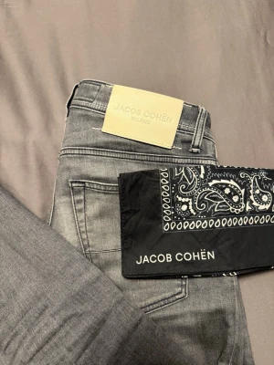 Grå jeans från Jacob Cohën - Snygga grå jeans från Jacob Cohën med klassisk femficksmodell och diskret logga på myntfickan. Jeansen har en riktigt schysst mörk/ljusgrå. Tillverkade i mjukt denimtyg som ger en skön känsla. Perfekta för dig som gillar stilrena och moderna plagg. Meddela vid eventuella frågor!