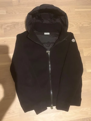 Svart stickad jacka från Moncler - Svart stickad jacka från Moncler med huva och dragkedja framtill. Jackan har Moncler-logga på ärmen och är fodrad på insidan av huvan. Perfekt för kyliga dagar och snygg till alla outfits.