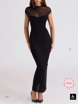 Svart långklänning med meshdetaljer - Elegant svart långklänning med transparent mesh upptill och korta ärmar. Klänningen har figurnära passform, hög hals och slits bakom. Perfekt för dig som vill ha en stilren och modern look.