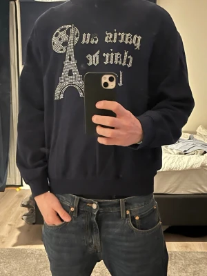 Mörkblå sweatshirt Paris-motiv - Säljer en mörkblå sweatshirt med ett glittrigt Paris-tryck och Eiffeltornet i strass på bröstet. Tröjan har rund halsringning och ribbade muddar vid ärmslut och nederkant. Perfekt för dig som gillar streetstyle och vill sticka ut lite extra.