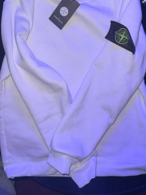 Vit hoodie från Stone Island - Säljer en fräsch vit hoodie från Stone Island med klassisk logga på ärmen. Hoodien har känguruficka framtill och ribbade muddar vid ärmslut och nederkant. Perfekt för dig som gillar streetwear och vill sticka ut med en ikonisk look.