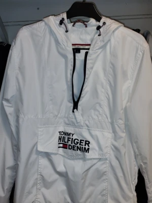 Ny Vit vindjacka Tommy Hilfiger Denim Medium - Snygg vit vindjacka från Tommy Hilfiger Denim med halv dragkedja framtill och stor ficka med logga på magen. Jackan har huva med dragsko och är tillverkad i lätt, vindtätt material. Perfekt för en sportig och stilren look. Ny. Normal passform.