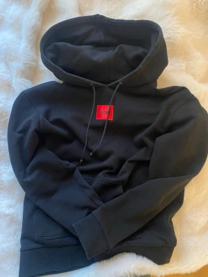 Svart hoodie från Hugo boss med röd logga - Snygg svart hoodie från Hugo boss med röd logga på bröstet. Hoodien har huva med dragsko, magficka och ribbade muddar vid ärmslut och nederkant. Tillverkad i mjukt bomullsmaterial. Unisex