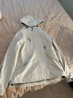 Vit vindjacka med huva - Snygg vit vindjacka med justerbar huva och dragkedjor framtill. Jackan har flera praktiska fickor med dragkedja och en modern, minimalistisk design. Perfekt för blåsiga dagar och enkel att matcha med olika outfits.