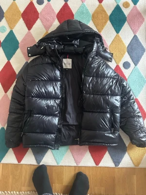 Svart dunjacka med huva från Moncler - Säljer en svart, glansig dunjacka från Moncler med avtagbar huva och två dragkedjeförsedda fickor. Jackan har klassisk Moncler-logga på ärmen och quiltad design som ger en riktigt puffig look. Perfekt för kalla vinterdagar.