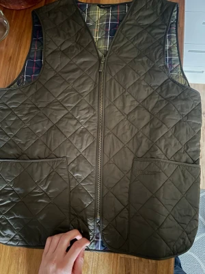 Barbour väst - Snygg olivgrön dunväst från Barbour med klassiskt rutigt innerfoder. Västen har två stora framfickor, dragkedja framtill och quiltat mönster. Perfekt lager på lager-plagg för kyliga dagar och ger en tidlös look. Kom privat för frågor eller mer bilder! Pris kan diskuteras