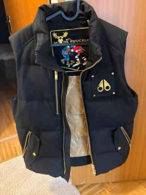 Svart dunväst från Moose Knuckles - Snygg svart dunväst från Moose Knuckles med guldfärgade dragkedjor och detaljer. Insidan har ett lyxigt guldmönstrat foder och en innerficka med dragkedja. Västen har hög krage och ikonisk logga på bröstet. Perfekt för lager-på-lager och streetwear.  Köptes ny förra året !!