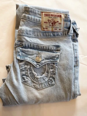 Ljusblå True Religion jeans - Säljer ett par ljusblå jeans från True Religion med ikoniska bakfickor och snygga slitningar. Jeansen har utsvängda ben, silverfärgade knappar och slitna bakfickor med klassisk design💓Stl 23, dvs ca Xs-S. Har lagad defekt på baksidan, se skick i bilder