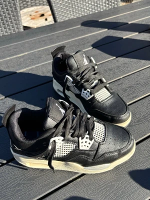 Svarta Air Jordan 4 sneakers - Säljer ett par svarta Air Jordan 4 sneakers med vita detaljer och meshpaneler. Skorna har klassisk siluett, svart läder och textil, samt Jumpman-logga på plösen. Storlek 37