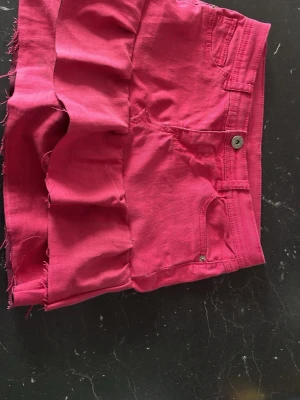 Rosa jeanskjol med volanger  - Säljer ett par rosa jeansshorts med rå kant och volangdetalj nedtill. Shortsen har klassisk femficksdesign, knapp och dragkedja framtill. Perfekta för sommardagar och ger en cool vibe till din outfit.har syr den själv av ett par jeans som var från Kappahl 