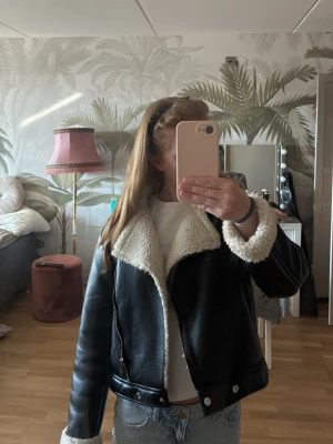Svart skinnjacka med fuskpäls - Snygg svart skinnjacka med vit fuskpäls på krage, ärmslut och insida. Jackan har en rak passform, dragkedja framtill och två knappar längst ner. Storleken som står passar inte in på hur den är igentligen enligt mig, storleken är 146-152 från Gina Young men den passar mig som bär XS, enligt mig är det snyggt med en kortare jacka nu till våren 🤗