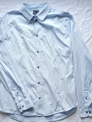 Ljusblå skjorta från H&M - Klassisk ljusblå skjorta från H&M med lång ärm och knappar framtill. Skjortan har en stilren krage och är tillverkad i mjuk bomull, perfekt för en clean och enkel look.
