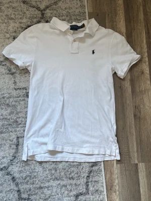 Vit pikétröja från Polo Ralph Lauren - Klassisk vit pikétröja från Polo Ralph Lauren med korta ärmar och den ikoniska broderade loggan på bröstet. Tröjan har krage, knappar framtill och är tillverkad i mjuk bomull. Perfekt till en clean och stilren look.
