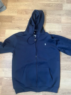 Marinblå hoodie från Polo Ralph Lauren - Snygg marinblå hoodie från Polo Ralph Lauren med dragkedja framtill, huva med dragsko och klassisk vit logga på bröstet. Mjuk bomullsblandning och två stora fickor framtill. Perfekt för en avslappnad och stilren look.