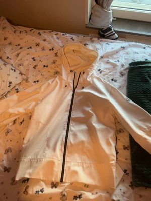 Vit regnjacka från Moncler XL - Snygg vit regnjacka från Moncler med huva och svart dragkedja. Jackan har minimalistisk design, justerbar huva och är tillverkad i vattenavvisande material med meshfoder för extra komfort. Perfekt för regniga dagar när du vill hålla stilen.