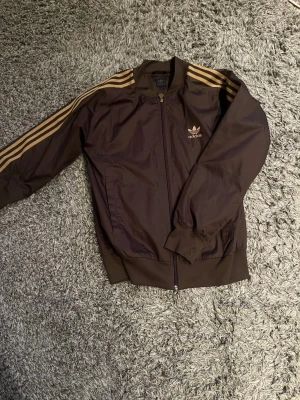 Brun vindjacka från Adidas - Snygg brun vindjacka från Adidas med klassiska guldfärgade ränder längs ärmarna och Adidas-logga på bröstet. Jackan har dragkedja framtill, ribbade muddar och krage. Perfekt för en sportig och retro look.
