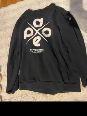 Svart sweatshirt från Dope - Säljer en svart sweatshirt från Dope med stort vitt tryck på bröstet där det står 'dope authentic sportwear'. Tröjan har rund halsringning och ribbade muddar vid ärmslut och nederkant. Perfekt för en avslappnad streetwear-look.