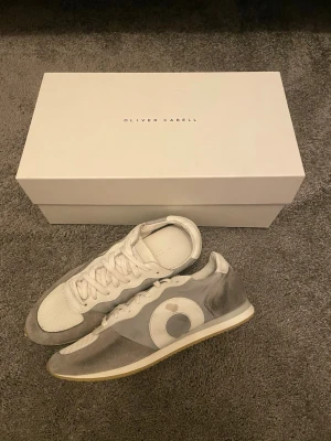 Gråvita runners från Oliver Cabell - Snygga runners från Oliver Cabell i grått och vitt med mesh och mockadetaljer. Skorna har en låg silhuett, vita snören och en gummisula i beige. Båda har en synlig defekt då det har skrapats av lite färg från cirkeln och är på vissa ställen i en lite mörkare nyans.