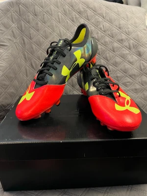 Under Armour fotbollsskor - Snygga fotbollsskor från Under Armour i rött och svart med stora gula loggor på sidorna. Skorna har snörning och är tillverkade i syntetmaterial för bra grepp och lätt känsla på planen. Perfekta för dig som vill sticka ut på fotbollsplanen.