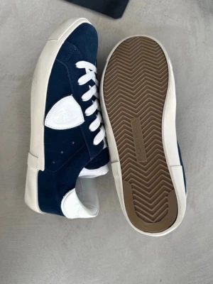 Philippe model skor - Snygga sneakers från Philippe model i mörkblå mocka med vita detaljer och vita skosnören. Skorna har en låg silhuett och en vit gummisula med fiskbensmönster undertill. Klassisk logga på sidan och bekväm innersula. Perfekta för en stilren och avslappnad look.