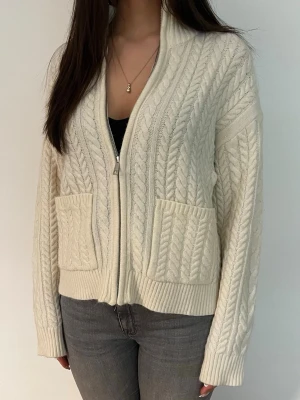 Vit kabelstickad kofta med dragkedja - Säljer en krämvit kabelstickad kofta med dragkedja framtill och två stora fickor. Koftan har ribbade muddar och hög krage som kan bäras både öppen och stängd. Perfekt för lager på lager och riktigt mysig till jeans. Använd två gånger