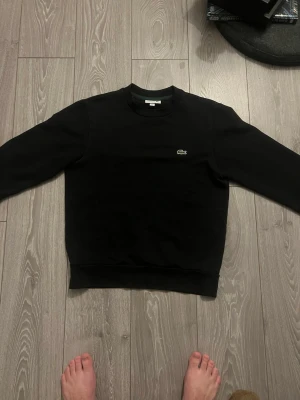 Svart sweatshirt från Lacoste - Klassisk svart sweatshirt från Lacoste med rund halsringning och den ikoniska gröna krokodilloggan broderad på bröstet. Tröjan har ribbade muddar vid ärmslut och nederkant. Perfekt för en clean och stilren look. Pris kan diskuteras 