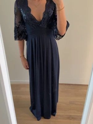 Marinblå långklänning - Elegant marinblå långklänning med transparenta trekvartsärmar och paljettbroderier upptill. Djup v-ringning och markerad midja ger en snygg siluett. Kjolen faller mjukt och materialet känns lyxigt. Perfekt för speciella tillfällen.