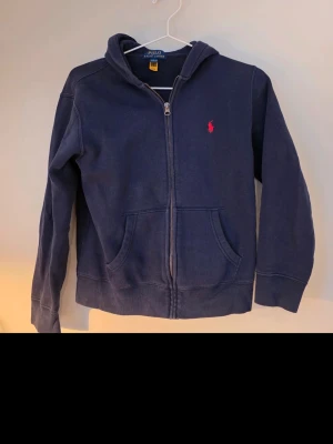 Mörkblå hoodie från Polo Ralph Lauren - Klassisk mörkblå hoodie från Polo Ralph Lauren med dragkedja framtill och två fickor. Ikoniska röda loggan broderad på bröstet. Tillverkad i mjukt bomullsmaterial och har en skön huva. Perfekt för en avslappnad och stilren look.