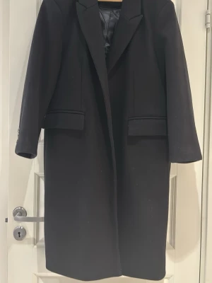 Svart klassisk ullkappa - Stilren svart kappa med rak passform och klassisk krage. Kappan har två fickor med lock framtill och knäppning vid ärmslut. Perfekt för en trendig och tidlös look. Fodrad insida för extra komfort.