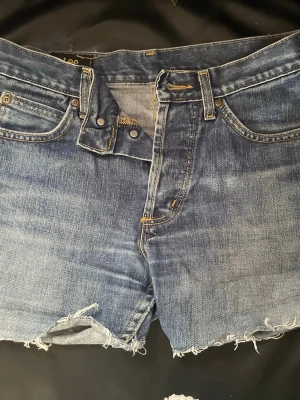 Blå jeansshorts från Lee - Snygga blå jeansshorts från Lee med råa, fransiga kanter och klassisk femficksdesign. Shortsen har normal passform och är tillverkade i slitstark bomull. Perfekta för sommardagar och har en cool, avslappnad look.