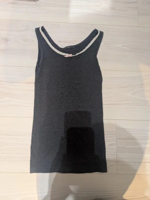 Mörkgrått/blått ribbat linne med spets - Mörkgrått/blått ribbat linne med breda axelband, dekorerat med vit spetskant runt halsringningen och en liten ljusrosa rosett framtill. Perfekt för mysiga hemmakvällar eller som lager under en tröja. Materialet känns mjukt och stretchigt. Har fått av min kusin så vet inte vart den är ifrån eller storlek men jag skulle säga XXS.