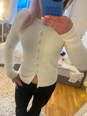 Vit kofta med pärlknappar ifrån Zara  - Snygg vit kofta med långa ärmar och dekorativa pärlknappar framtill. Koftan är ribbstickad vid ärmslut och nederkant, vilket ger en skön passform. Perfekt att styla till jeans eller kjol för en clean look.