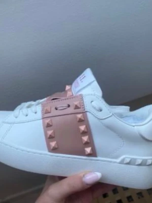 Valentino skor  - Snygga vita sneakers från Valentino med en bred rosa rem över sidan dekorerad med pyramidformade nitar. Skorna har en klassisk låg siluett, vita snören och detaljerade sömmar. Tillverkade i slätt läder för en clean och exklusiv look.