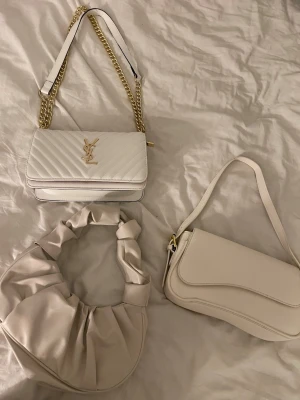 Vit YSL handväska med guldkedja - Säljer en snygg vit handväska från YSL med guldfärgad kedja och klassisk YSL-logga framtill. Väskan har quiltat mönster och axelrem i både läder och metall. Perfekt för dig som vill ha en trendig och lyxig accessoar till din outfit.Och dom andra två väskorna