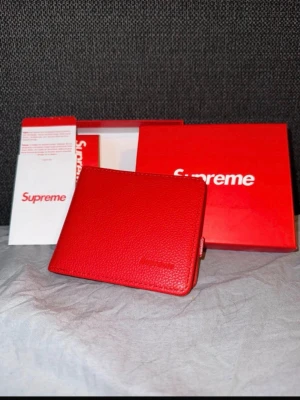 Röd plånbok från Supreme - Supreme plånbok i starkt rött konstläder med präglad logga på framsidan. Insidan har flera kortfack och ett myntfack med knappstängning. Kommer med originalförpackning och Supreme-kort. Perfekt för dig som gillar streetwear och vill ha något unikt.