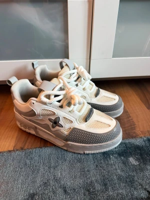 Vita och grå sneakers med stjärndetalj - Snygga sneakers i vitt och grått från Bape Sta med cool stjärndetalj på sidan. Skorna har chunky sula, meshpaneler och breda vita skosnören. Perfekta för dig som gillar streetwear och vill sticka ut med en unik look.