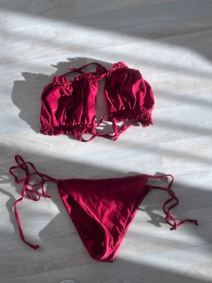 Mörkröd bikini med snörning - Trendig mörkröd bikini med topp som har volangdetaljer och knyt i nacken. Bikinitrosan har snörning i sidorna för justerbar passform. Perfekt för sommardagar på stranden eller vid poolen.