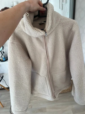 Beige teddyjacka från 157 - Mysig beige teddyjacka från 157 med hög krage och dragkedja framtill. Jackan har två stora fickor och är gjord i fluffigt teddy-material som håller dig varm. Perfekt för kyliga dagar och enkel att matcha med olika outfits.