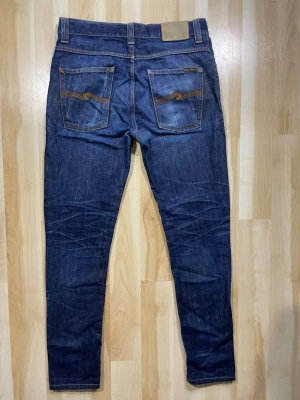 Mörkblå jeans från Nudie Jeans - Snygga mörkblå jeans från Nudie Jeans med klassisk femficksdesign och kontrastsömmar på bakfickorna. Jeansen har en smal passform och är tillverkade i denim av bomull. Perfekta för en stilren och avslappnad look.