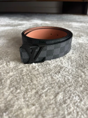 Fint bälte - Snyggt bälte  med klassiskt rutigt mönster i svart och grått. Bältet har en stor logga som spänne och insidan är beige. Perfekt accessoar för att lyfta din outfit.